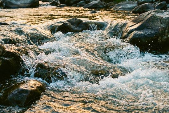 Água de rio fluindo com força entre pedras sob luz do sol, representando a relação natural entre Tantra e sexualidade e o fluxo de energia vital.