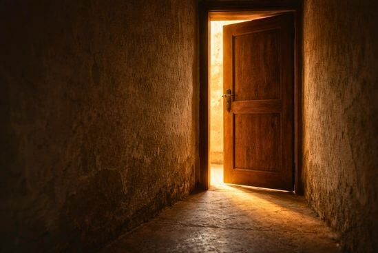 Porta de madeira escura entreaberta em corredor estreito de parede envelhecida, com faixa de luz quente entrando pelo vão e iluminando o chão — imagem de limiar que traduz a pergunta central sobre como saber se o trabalho é massagem tântrica ou terapia tântrica além do nome.