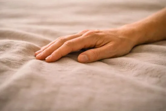 Mão adulta com a palma voltada para baixo sobre tecido de linho enrugado pelo peso dos dedos, com luz difusa de final de tarde, representando o como saber se um toque é bem-vindo quando o corpo finalmente encontra onde pousar.