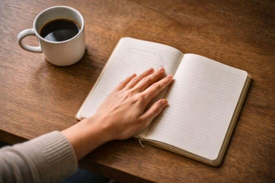 Mão com dedos pousados sobre página de caderno aberto em branco, ao lado de xícara pela metade em superfície de madeira com luz lateral — imagem do gesto de quem parou para observar pessoas que drenam sua energia antes de nomear o que percebe.