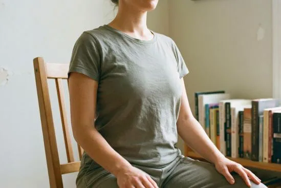 Torso relaxado de pessoa sentada em cadeira doméstica simples com luz natural lateral, ilustrando como o tantra no dia a dia se pratica em momentos comuns de atenção ao próprio corpo.