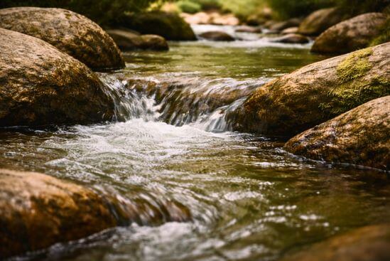 Corrente de água fluindo suavemente entre pedras de tom âmbar com luz refletida na superfície, simbolizando o retorno da vitalidade criativa e o prazer como fluxo que volta a correr após longa contenção.