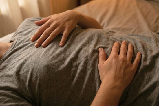 Duas mãos com peso real sobre peito e ventre de corpo em repouso com luz quente de quarto, evocando quando procurar apoio após a terapia tântrica como gesto de presença e cuidado consigo.