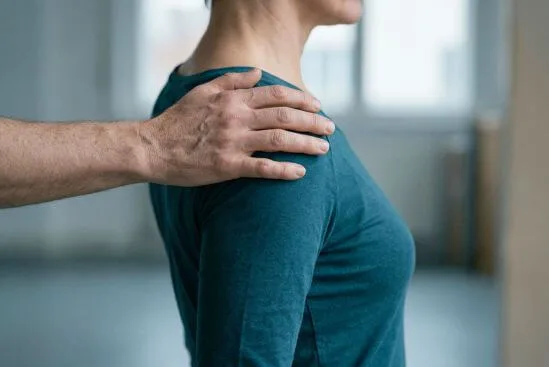 Mão adulta sobre o ombro de outra pessoa enquanto o tronco de quem recebe se inclina discretamente para o lado oposto, mostrando a diferença entre toque com consentimento e contato que o corpo tolera sem receber.